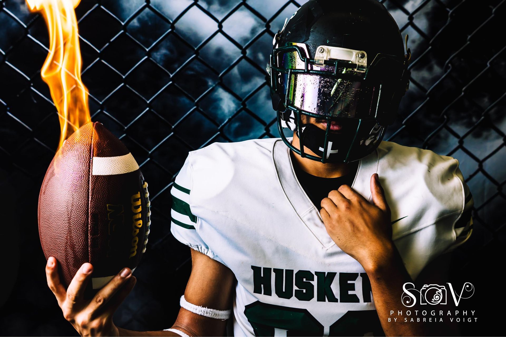 Football player in helmet and jersey with a burning football, against a chain-link fence and cloudy sky.