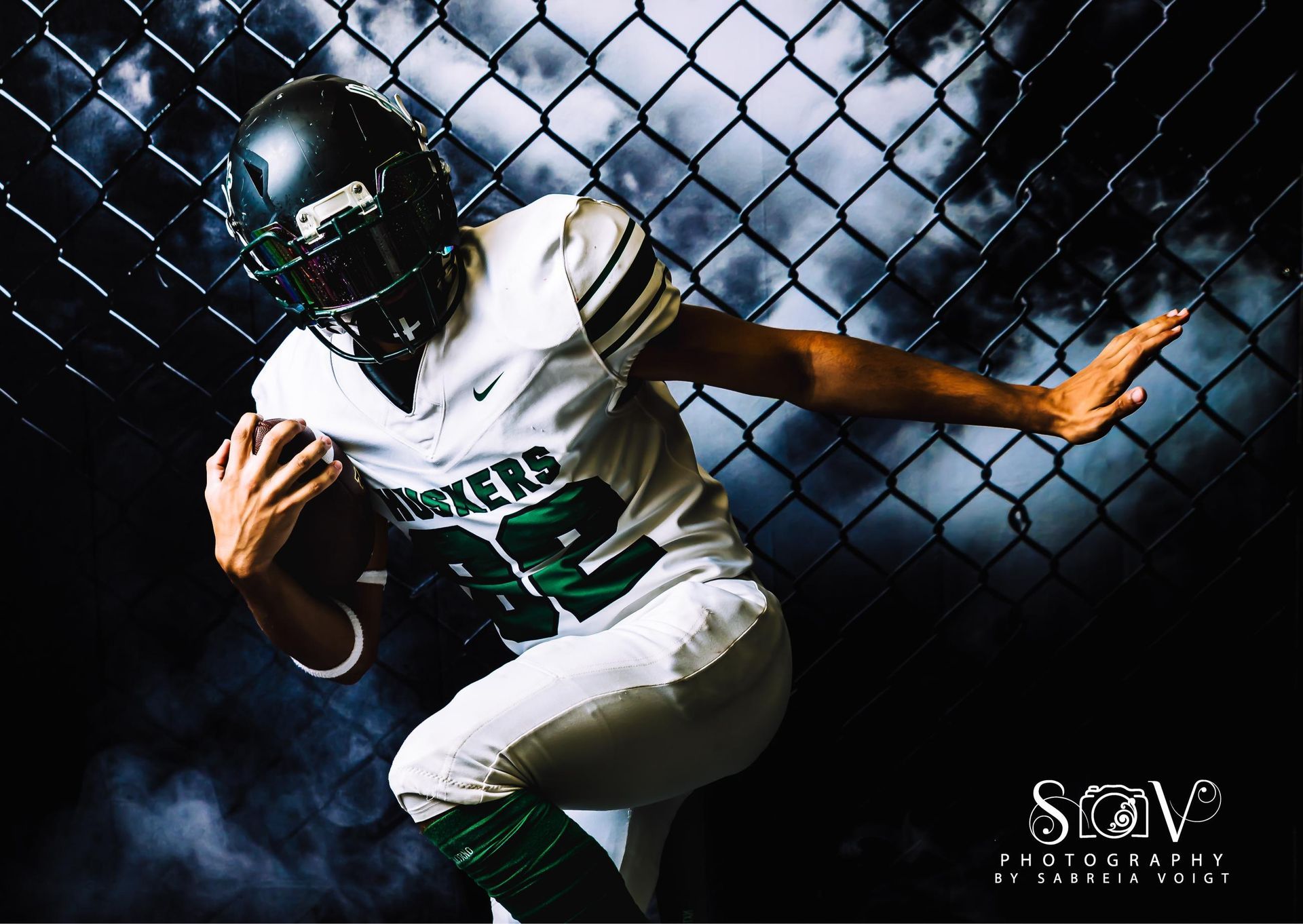Football player in white and green uniform runs with ball, against chain-link fence and smoky background.