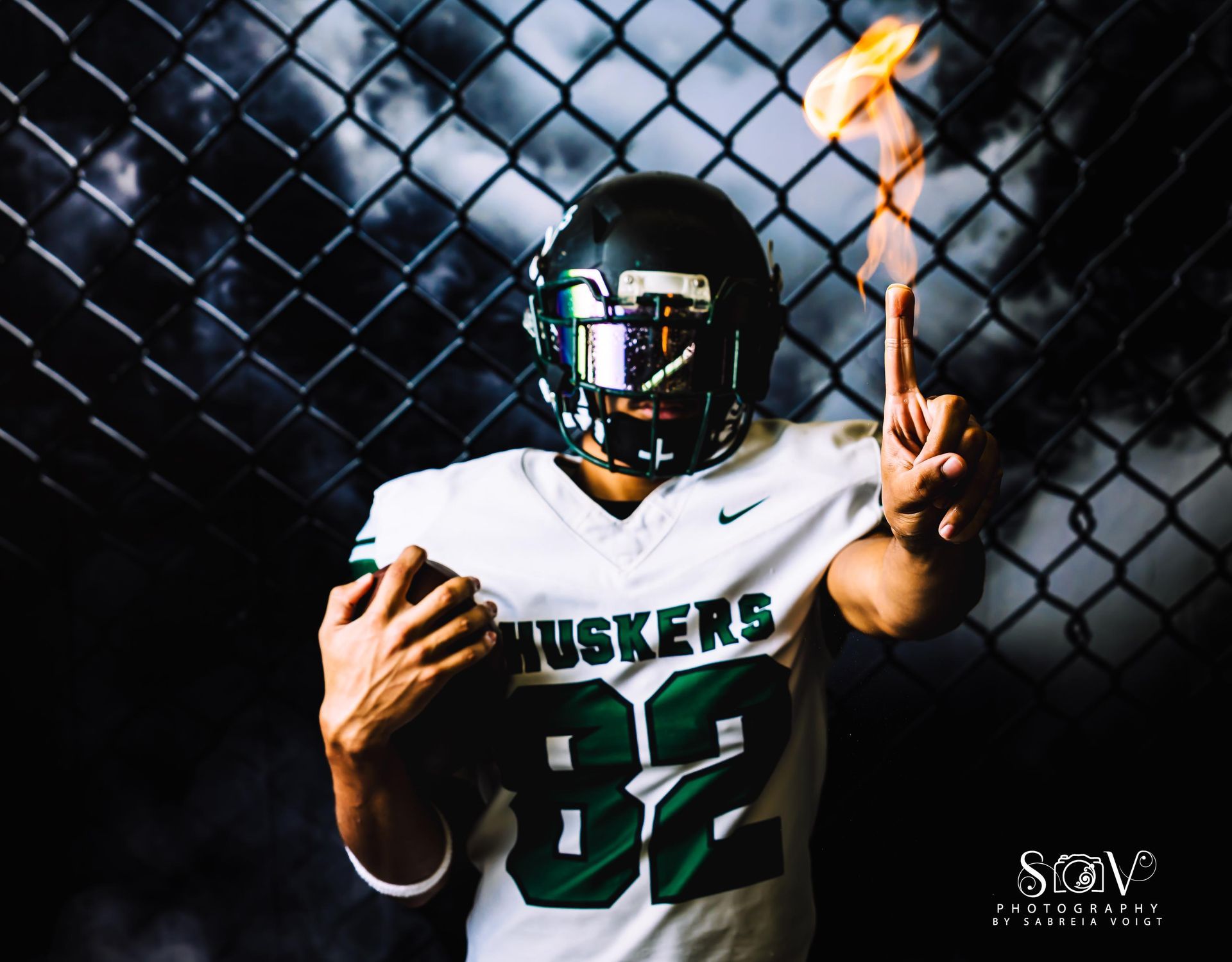 Football player in green and white jersey with helmet, holding ball, flames on finger, in front of chain-link fence.