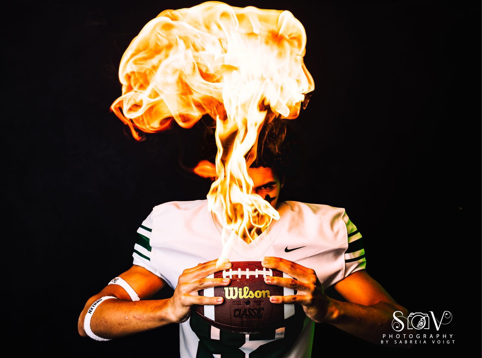 Football player holding football with flames erupting over it, black background.