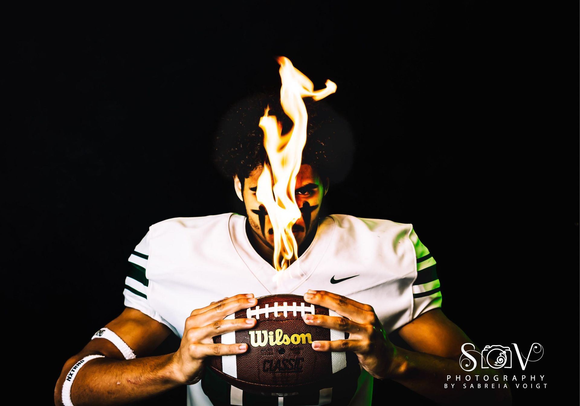 Football player holding flaming football. Wearing green and white jersey with black background.