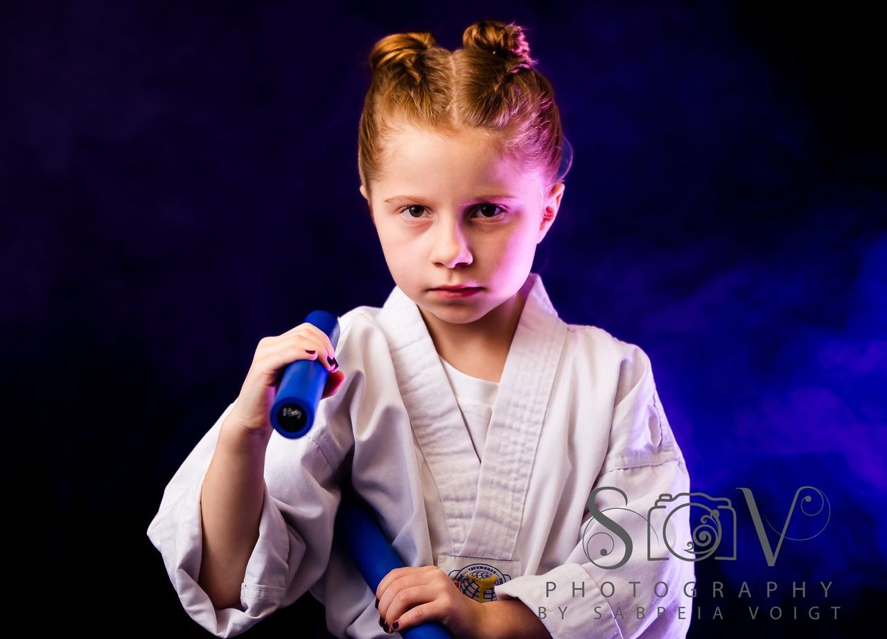 Girl in karate gi holding blue nunchucks, looking sternly at the viewer against a smoky purple and black background.