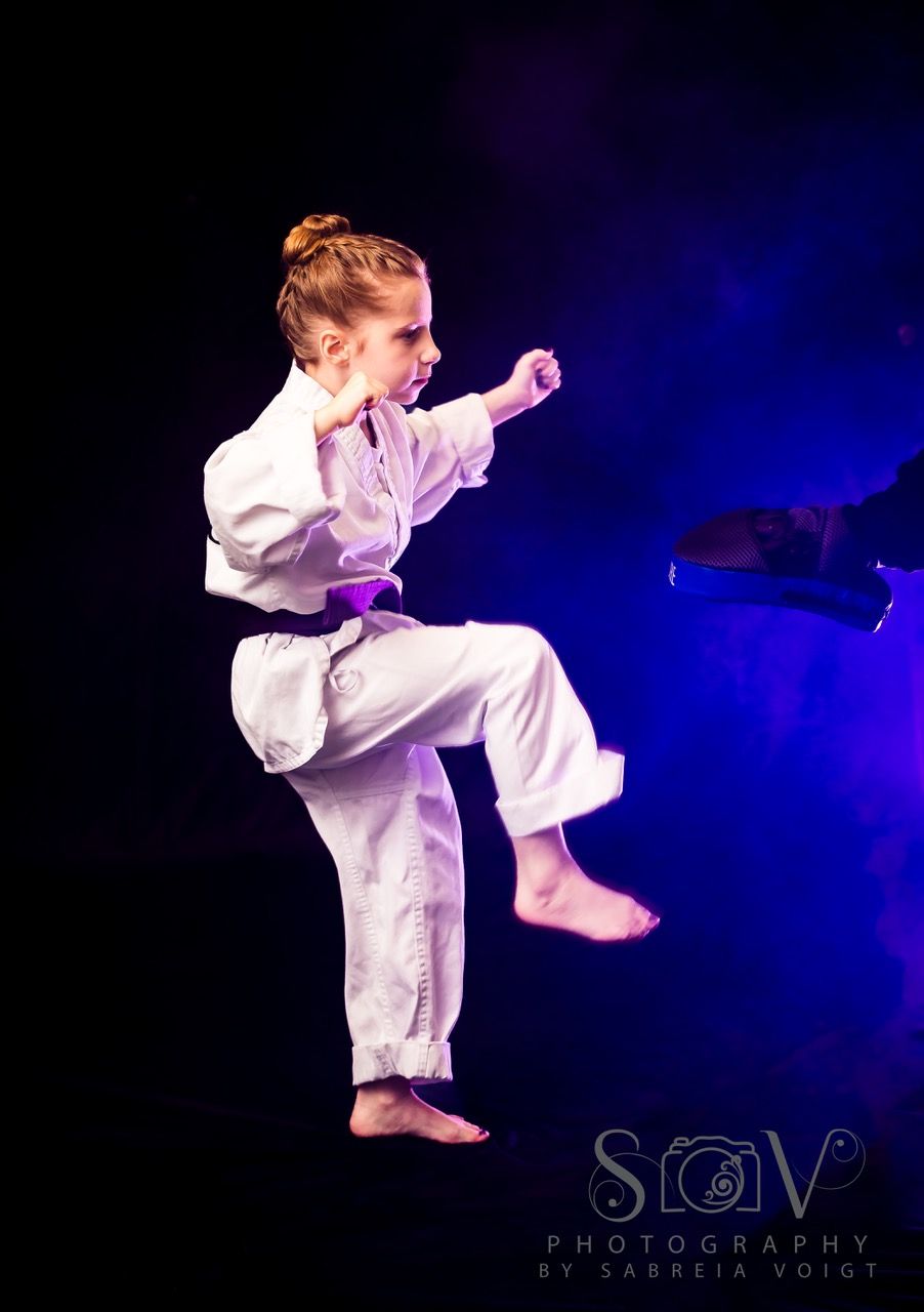 Young person in karate uniform, performing a kick. Dark background with blue lighting.
