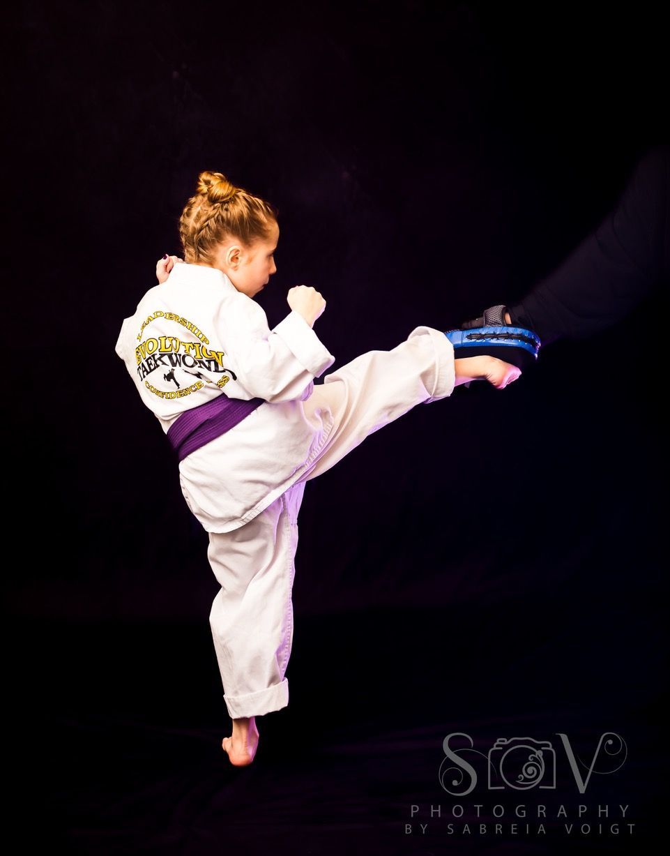 Young martial artist in white uniform, kicking a pad held by an arm against a black background.