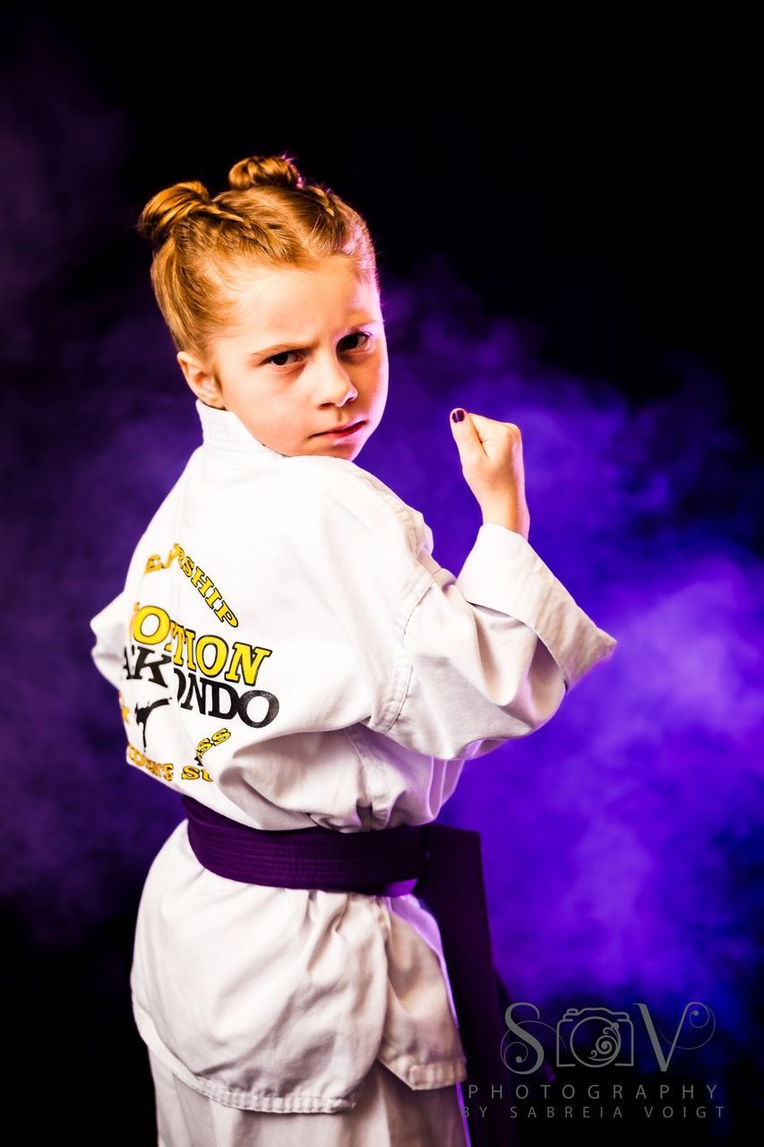 Girl in karate uniform with purple belt, fists raised, posing in front of purple and black background.