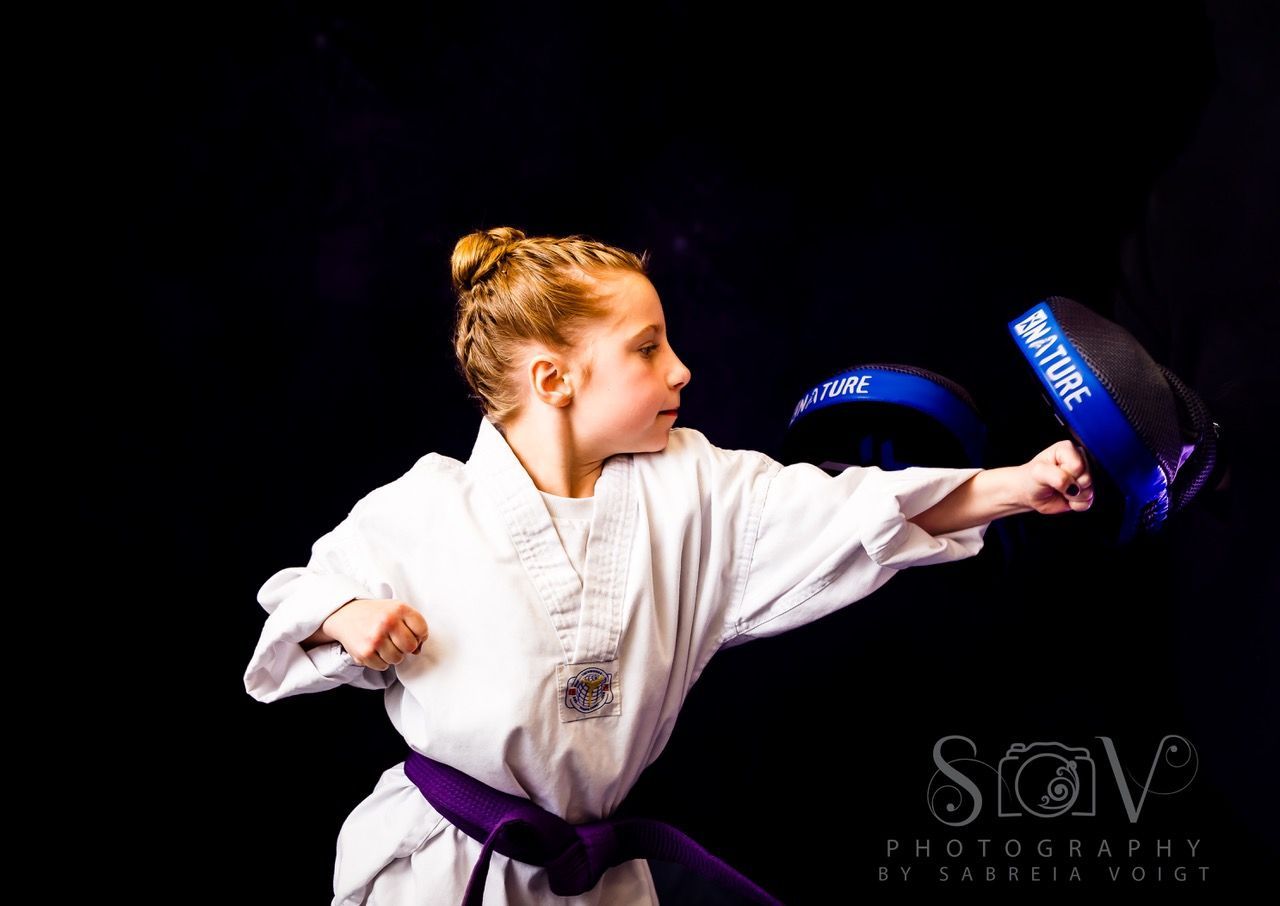 Young person in a martial arts uniform punches a pad, black background.
