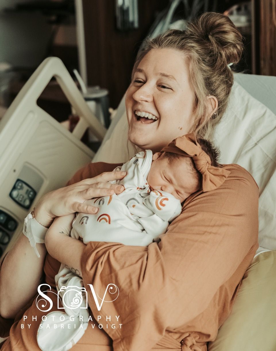 Woman smiles, cradling newborn in hospital bed. Baby wears rainbow onesie.
