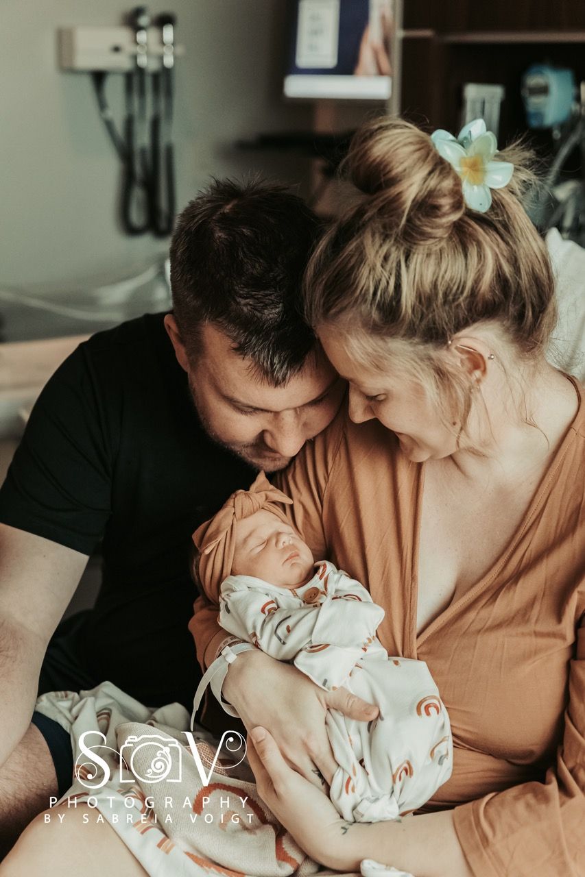 Parents cradling newborn baby in hospital room, baby wearing a headband and patterned onesie.