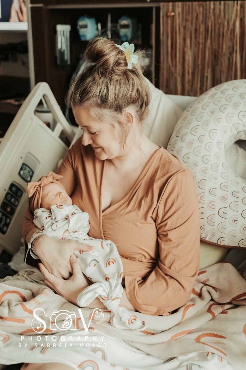 Woman holding newborn baby, smiling. Soft lighting, hospital room setting.