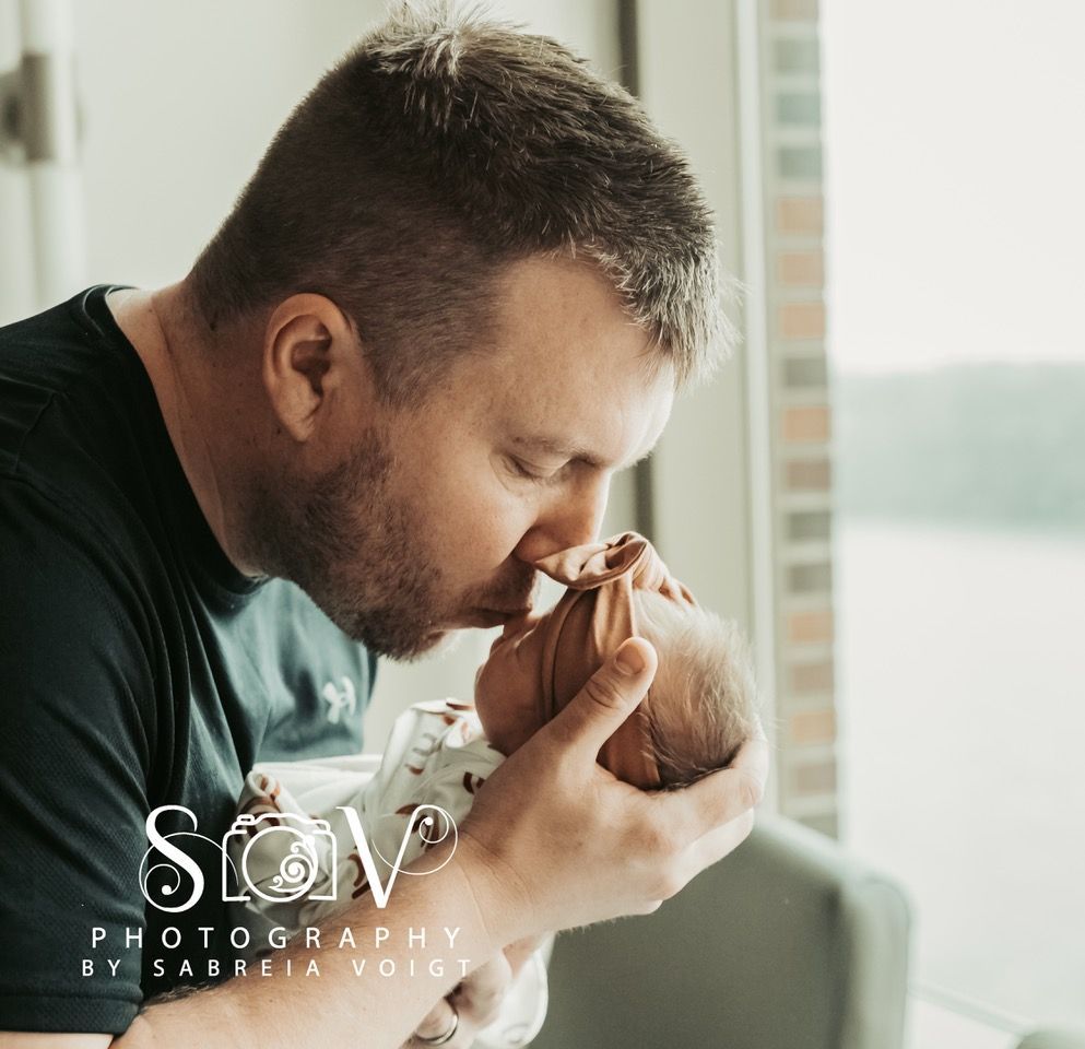 Man kissing a newborn baby's forehead. They are near a window, soft lighting.