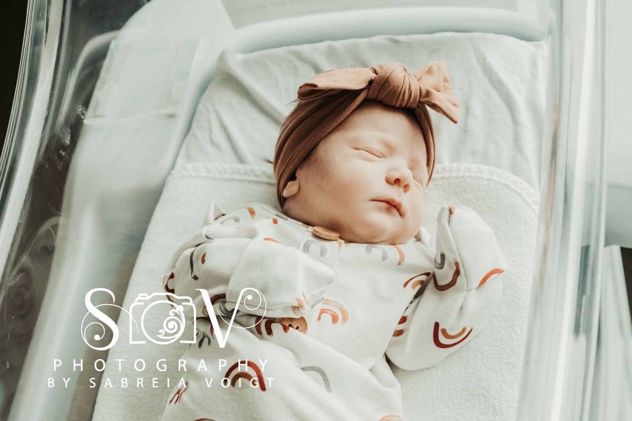 Newborn baby sleeping in a hospital bassinet, wearing a bow headband and rainbow-patterned onesie.