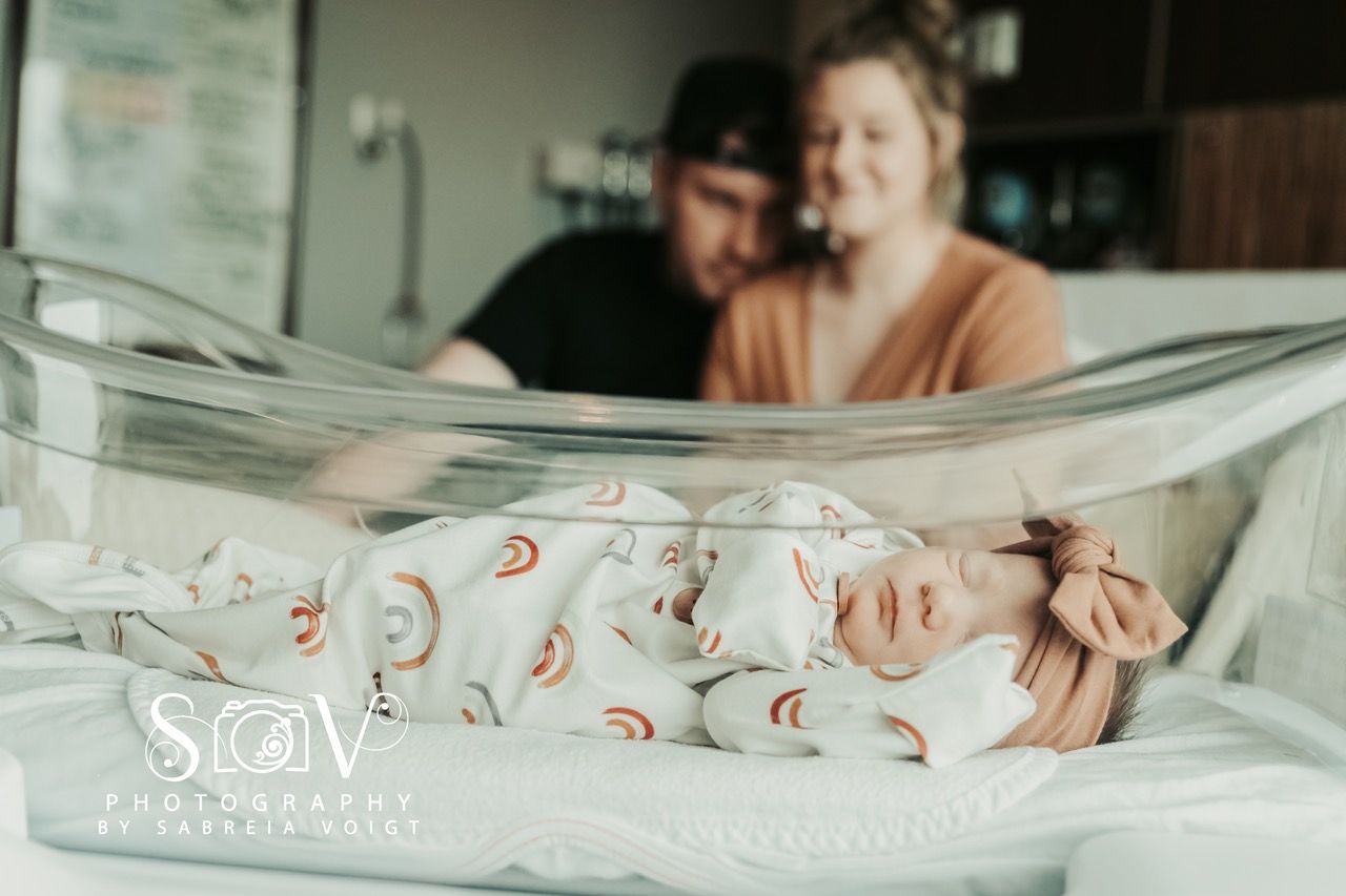 Newborn baby in bassinet, parents blurred in background, hospital setting.