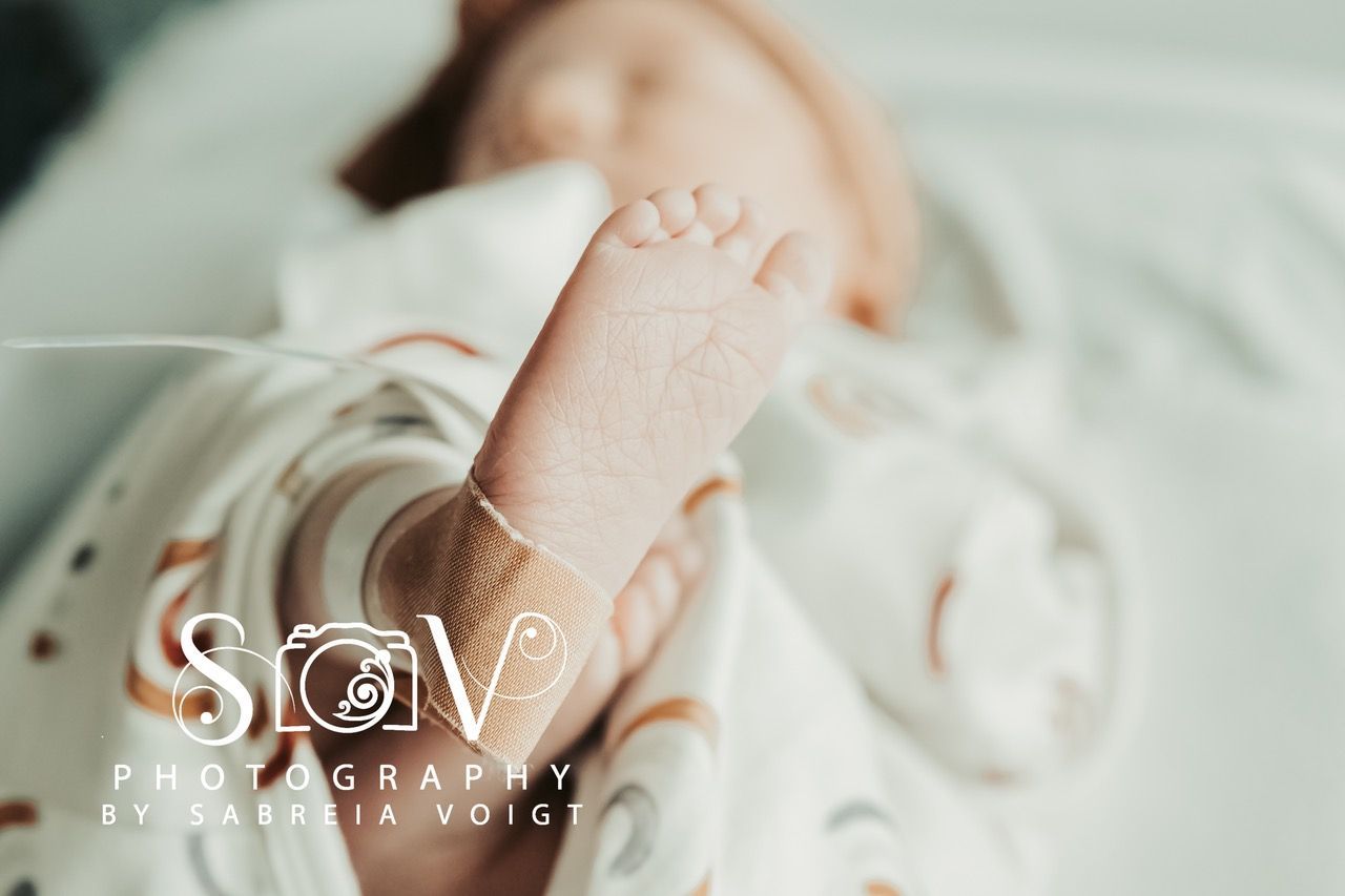 Close-up of a baby's foot with a medical sensor. Baby is wearing a rainbow-patterned onesie, on a white surface.