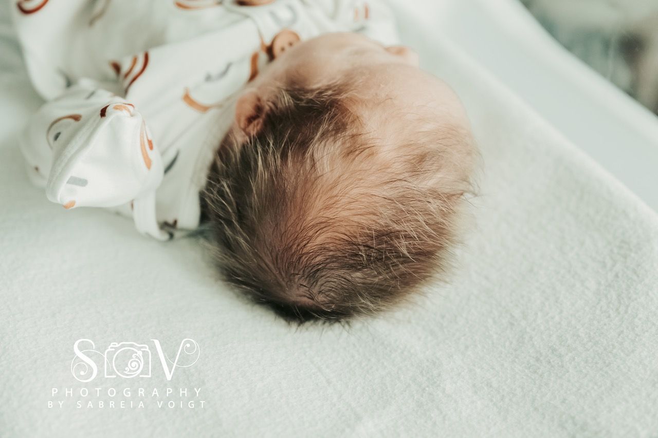 Close-up of a baby's head with wispy dark hair on a white surface, part of a rainbow-patterned onesie visible.