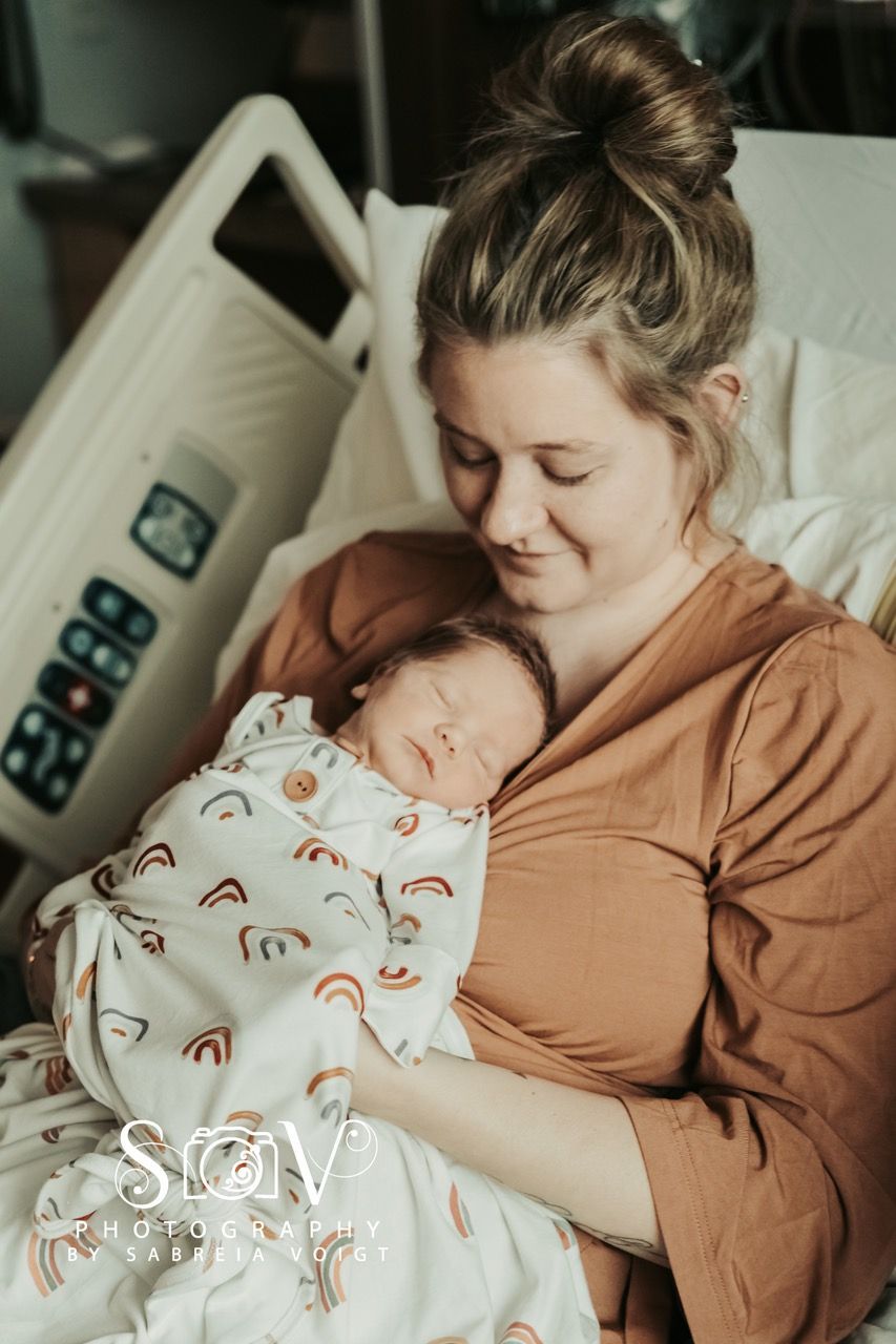 New mother holding newborn in hospital bed; baby wrapped in a blanket with rainbows, smiling mother.
