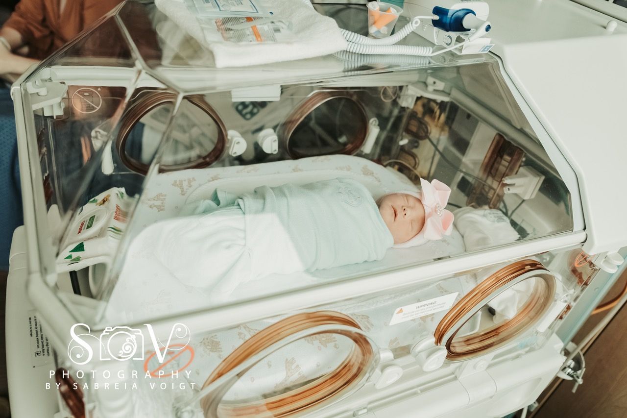 Newborn baby in an incubator, wearing a pink bow, in a hospital.