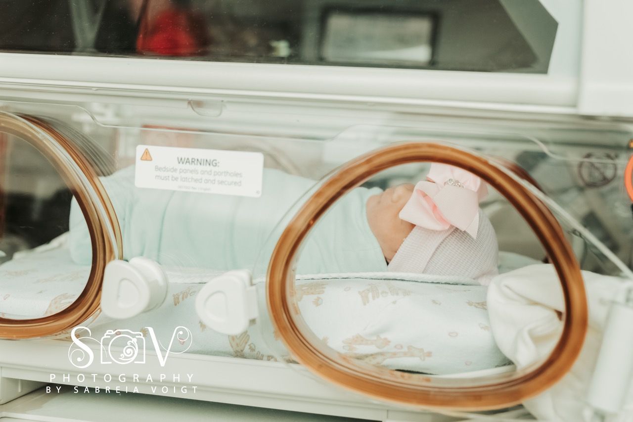 Newborn baby in an incubator, wearing a pink bow, in a hospital setting.
