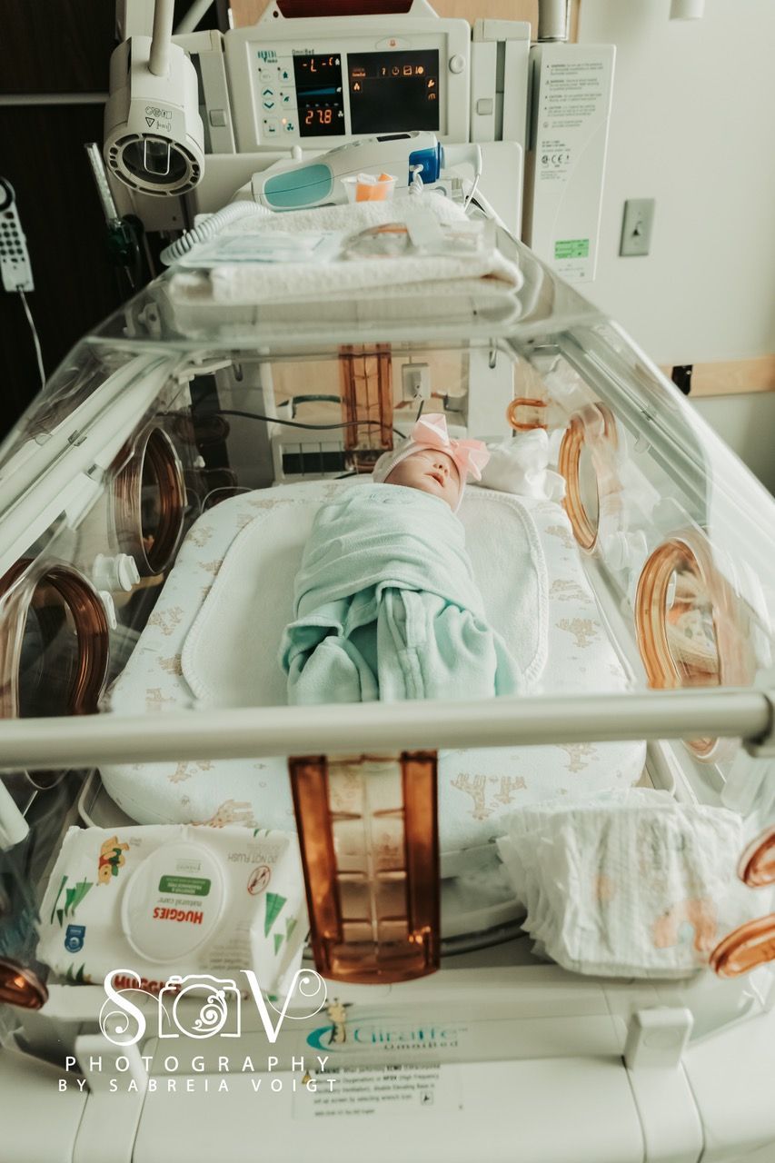 Baby swaddled in a hospital incubator, medical equipment visible, wipes and diapers on counter.