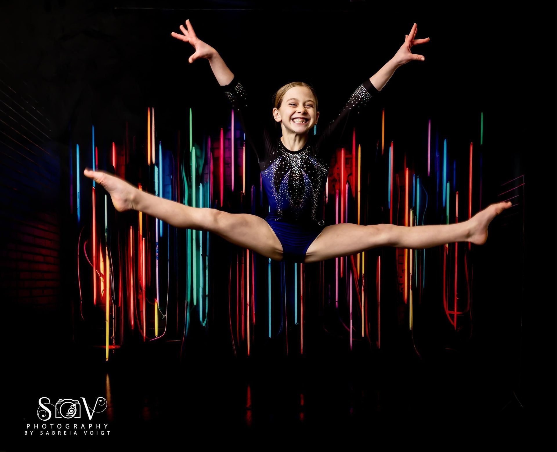 Gymnast doing a split leap against a backdrop of colorful lights; she is smiling with arms raised.