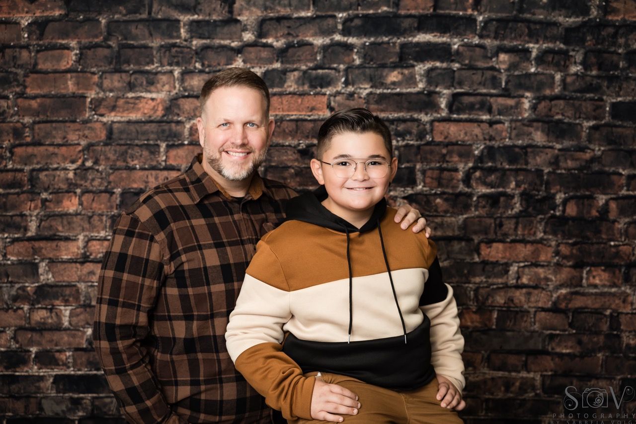 Man and boy pose together in front of a brick wall. Man has arm around boy.