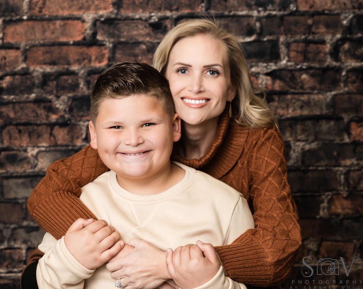 Woman hugging a smiling boy, both wearing sweaters, in front of a brick wall.