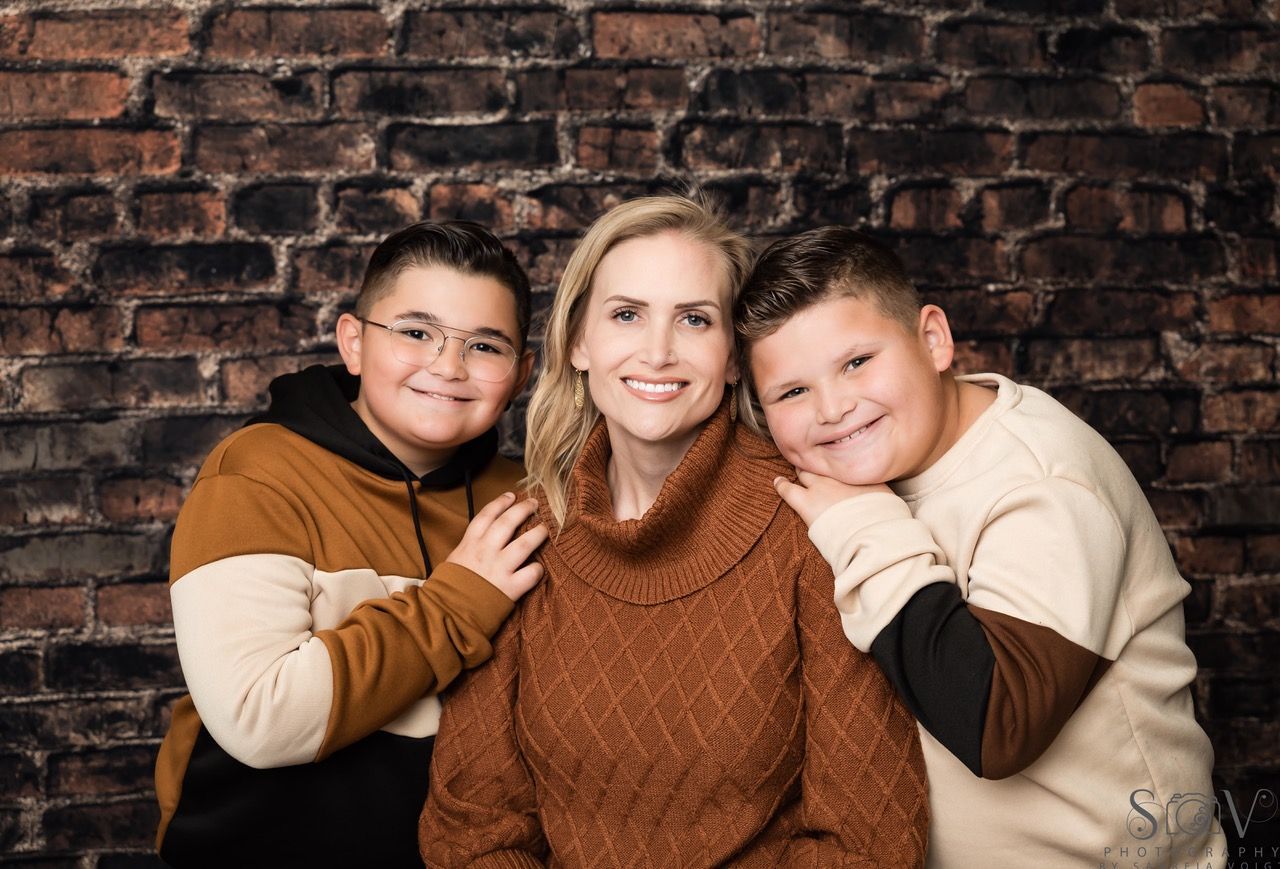 Woman and two children smiling in front of a brick wall. Woman wearing a brown sweater, boys in hoodies.