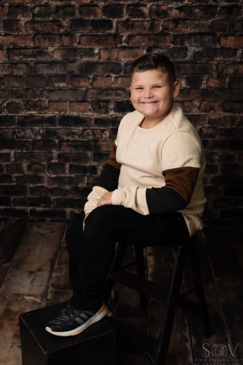 Boy in tan and brown shirt, black pants, and sneakers, smiles, sitting on stool in front of brick wall.