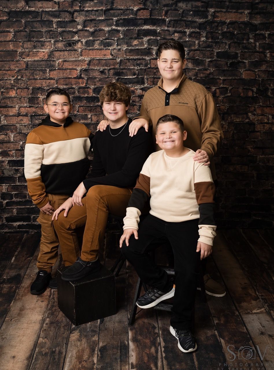 Four people posing in front of a brick wall. Three seated, wearing sweaters; one standing, wearing a brown sweater.