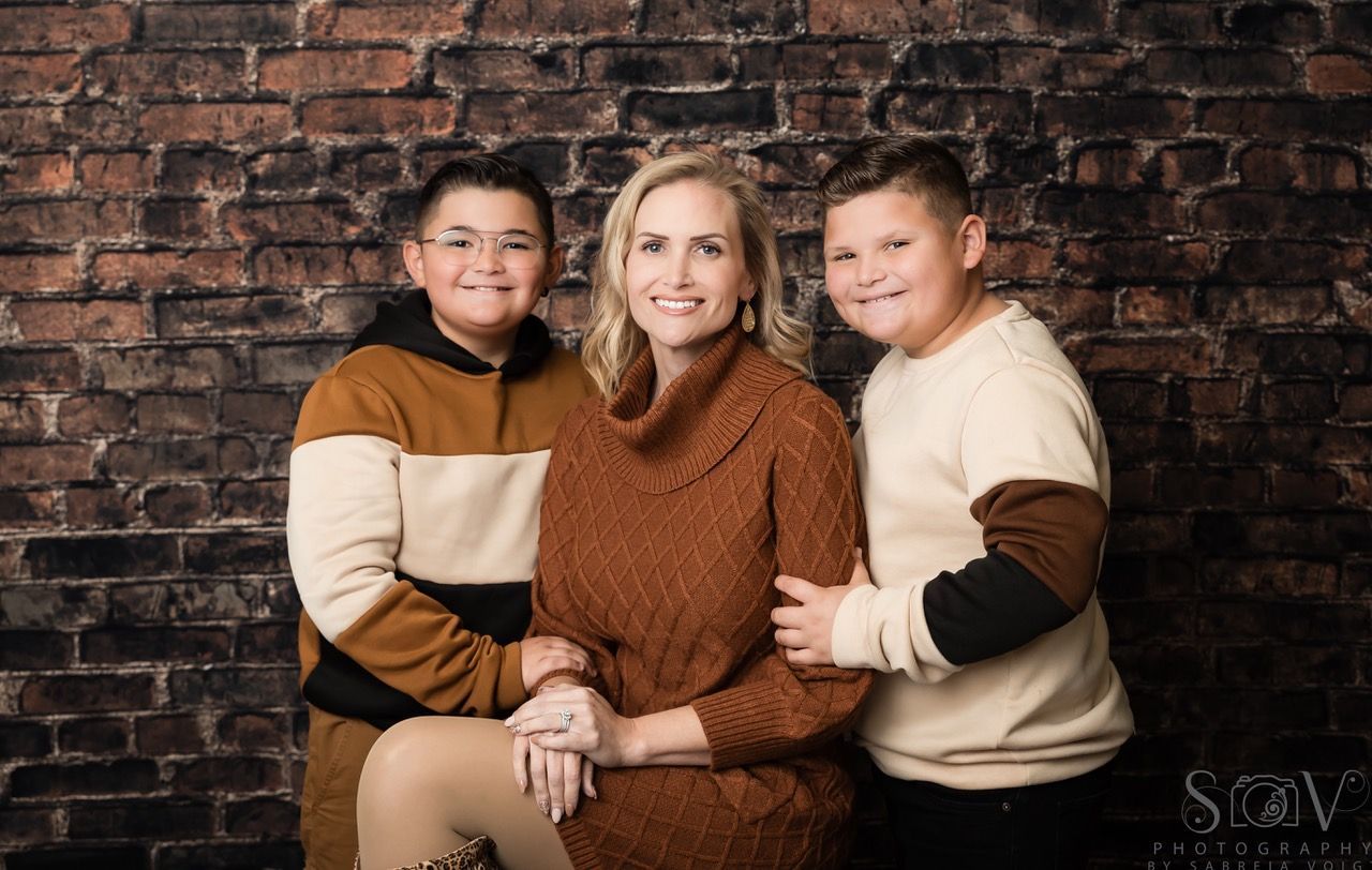 Woman and two children in front of a brick wall, all wearing earth-toned sweaters; smiling and posing.