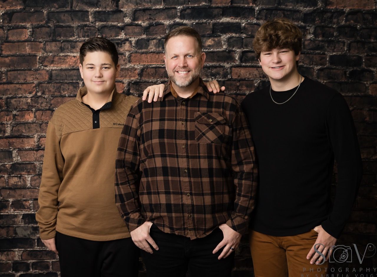 Man with two young people in front of a brick wall; man has arm around one.