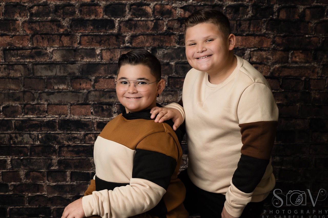 Two boys in sweaters pose in front of a brick wall, one with arm on the other's shoulder, both smiling.