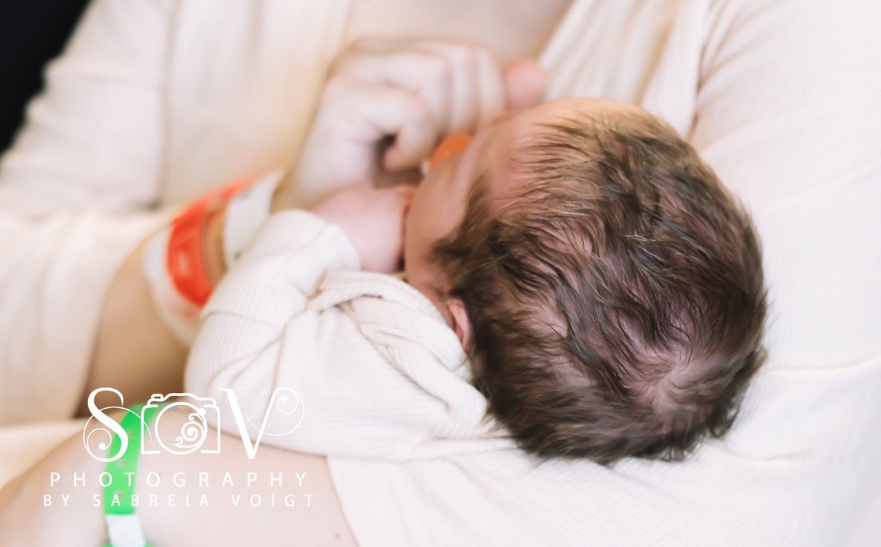 Newborn baby held in arms, wearing hospital wristbands. Dark hair, light-colored clothing.