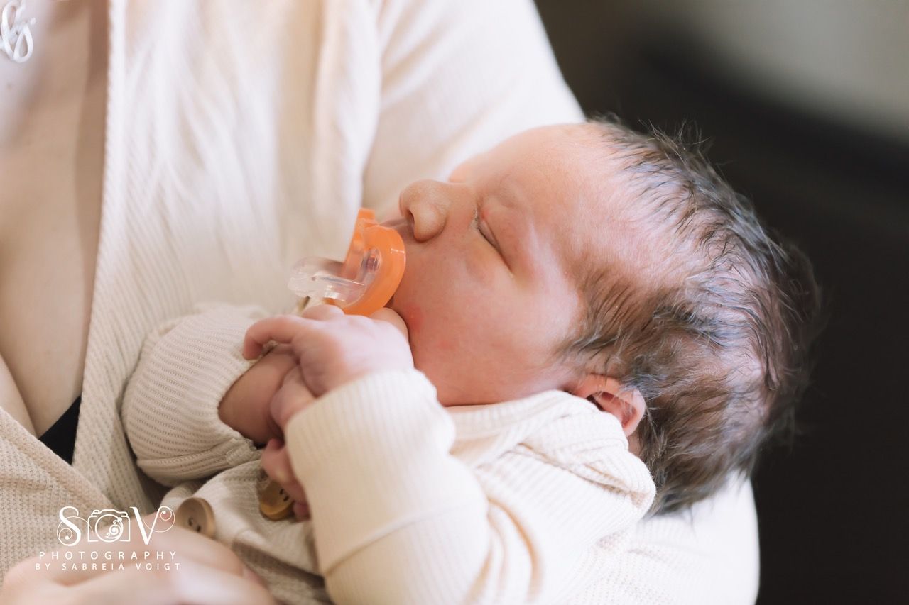 Newborn baby asleep, holding a pacifier, in someone's arms.