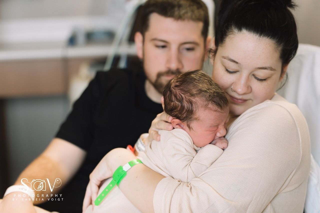 Newborn baby held by mother, father looks on; hospital setting.