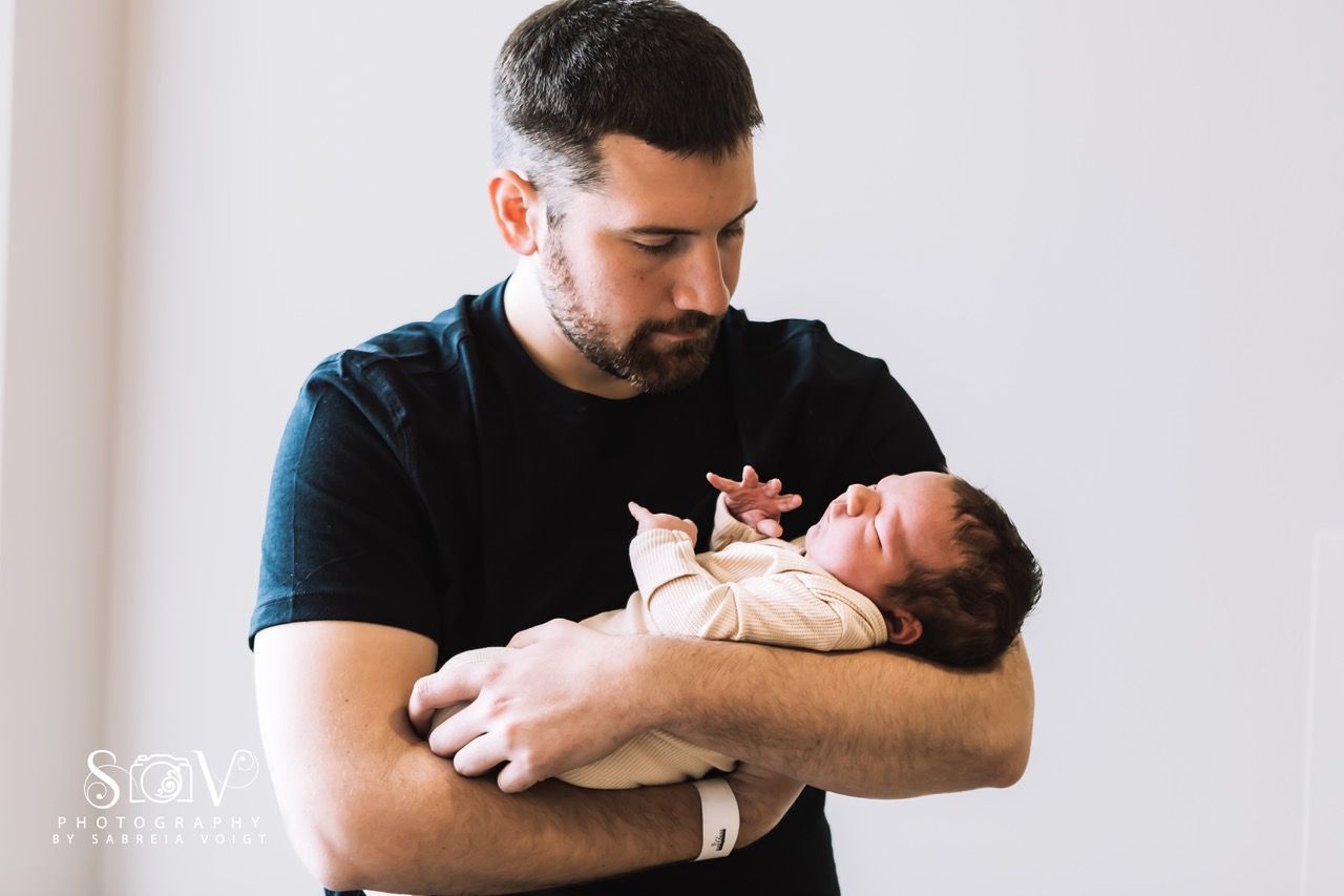 Man holding newborn baby, looking down with a gentle expression. White background.
