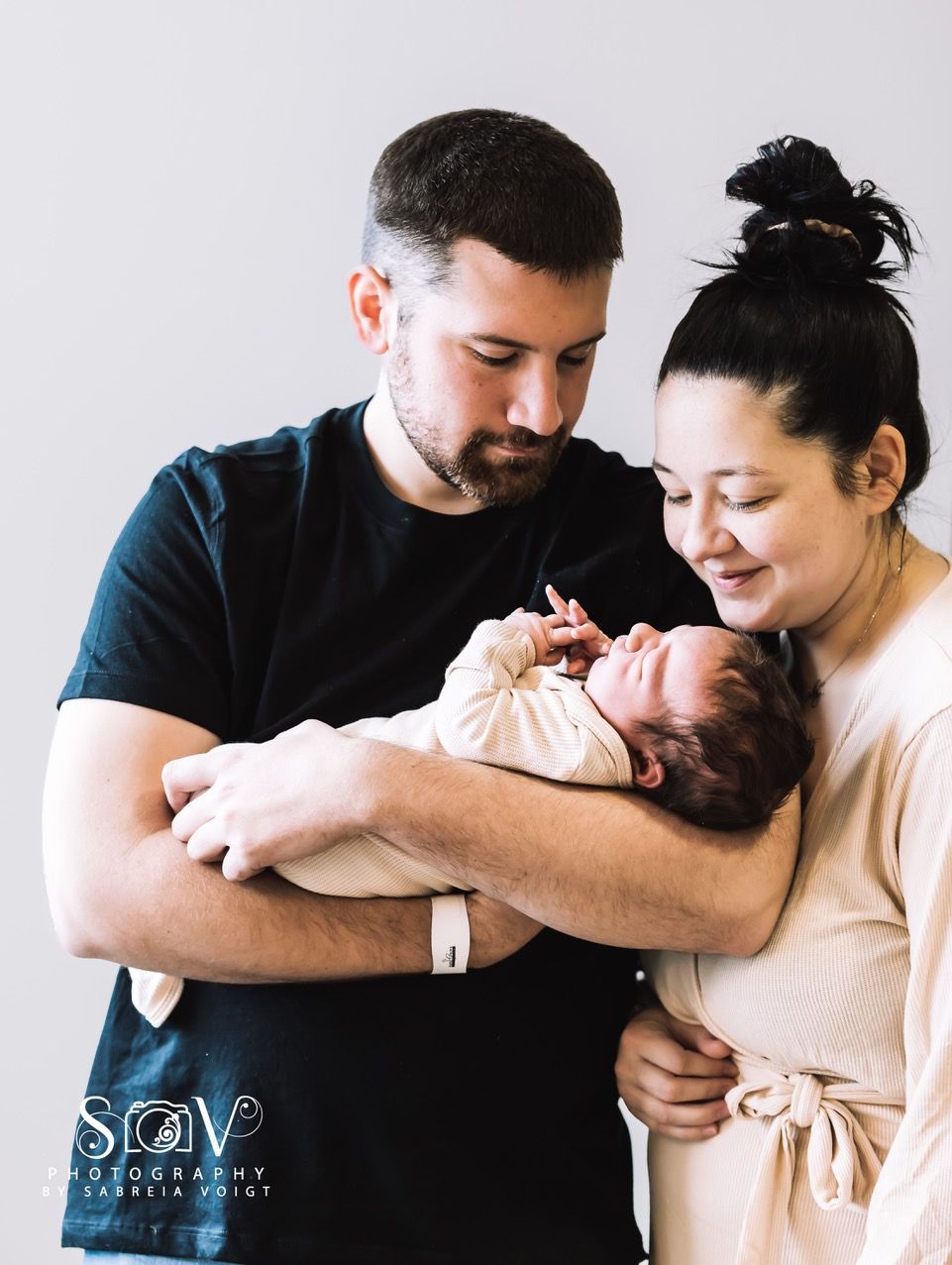 Parents gazing adoringly at newborn baby swaddled in arms. Light gray background.