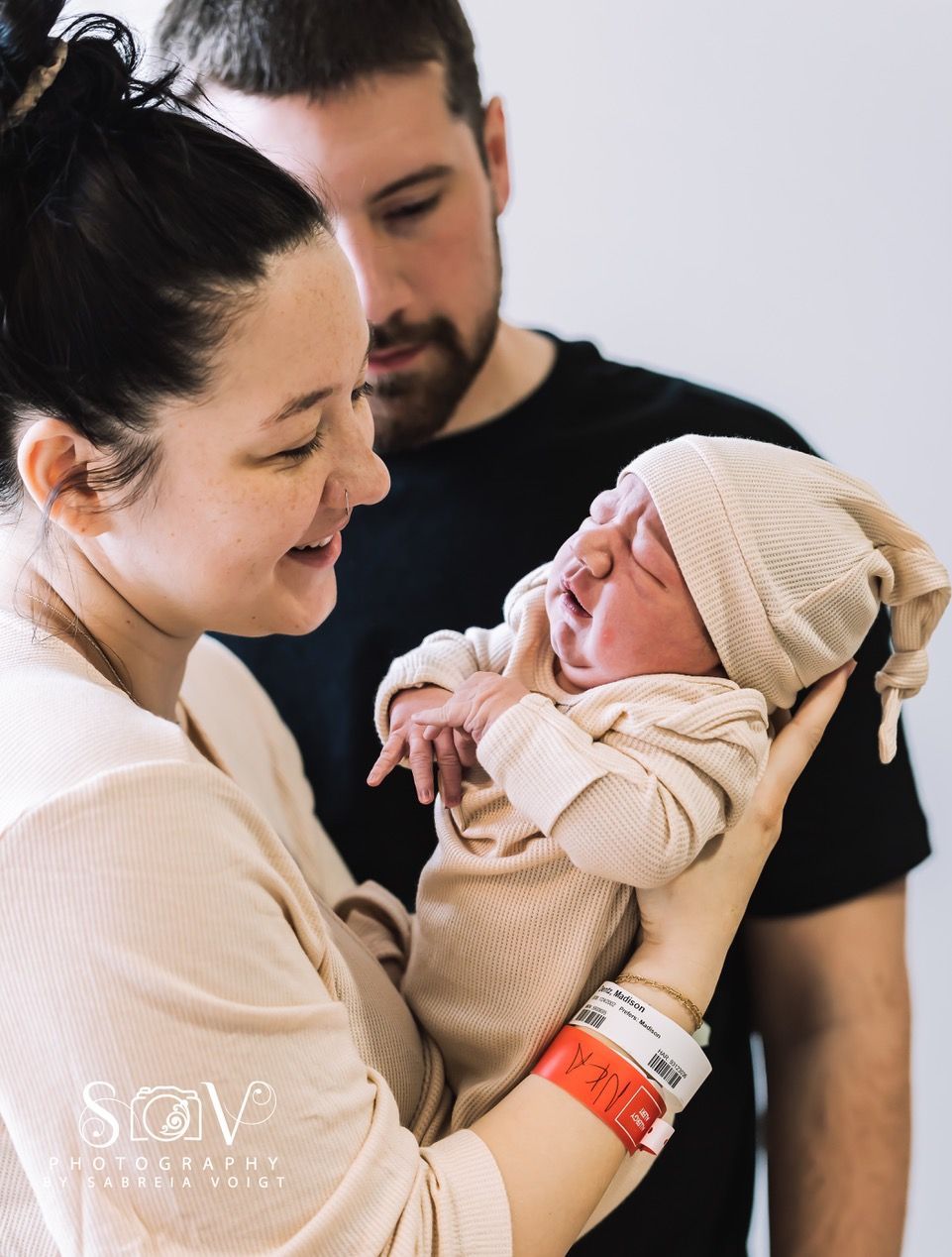 Parents gaze at newborn baby, held in arms. Baby wears a hat and light colored outfit.