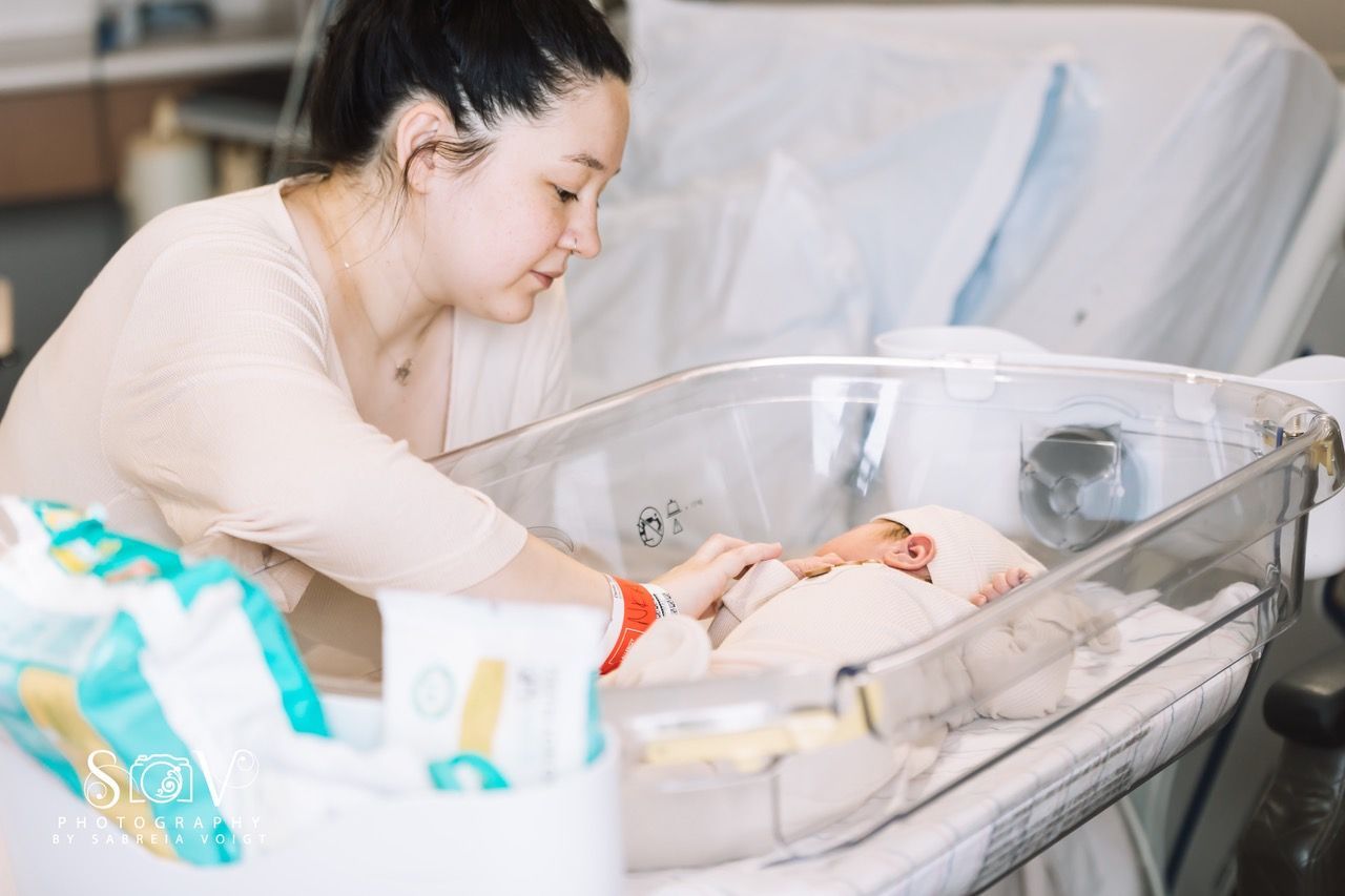 Woman leans over a hospital bassinet, looking at a newborn baby inside. White sheets and medical setting.