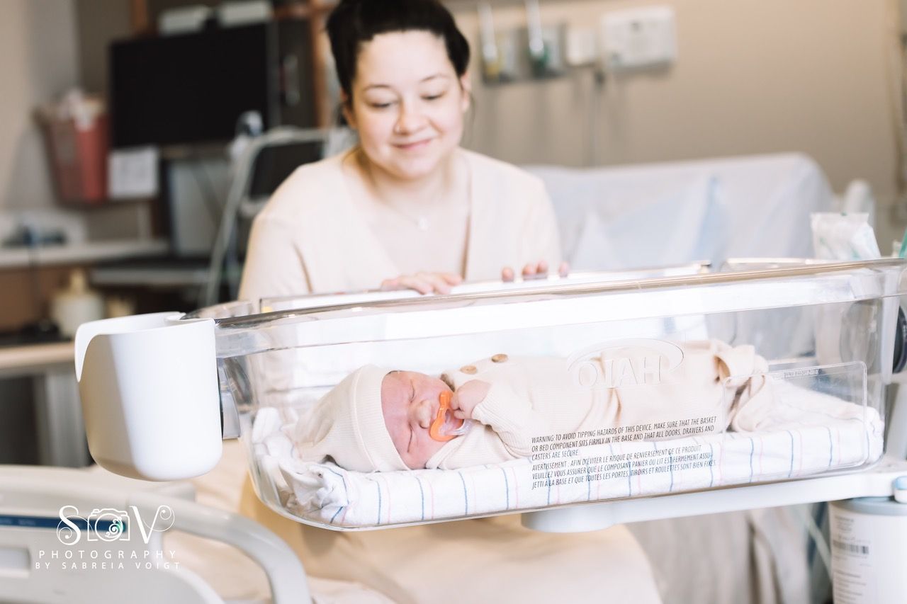 Newborn baby in clear hospital bassinet, mother smiling, hospital room setting.