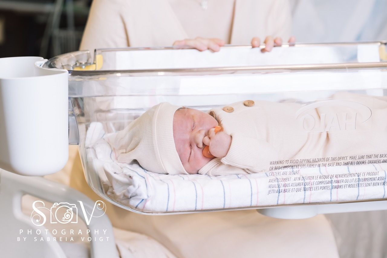 Newborn baby swaddled in hospital incubator, held by a person in a tan shirt.