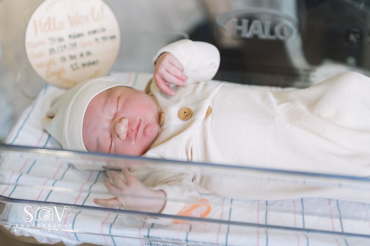 Newborn baby in a hospital bassinet, wearing a cream-colored outfit. A 