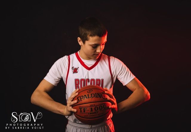Basketball player in white jersey, holding a ball, looking down. Red and black background with spotlights.