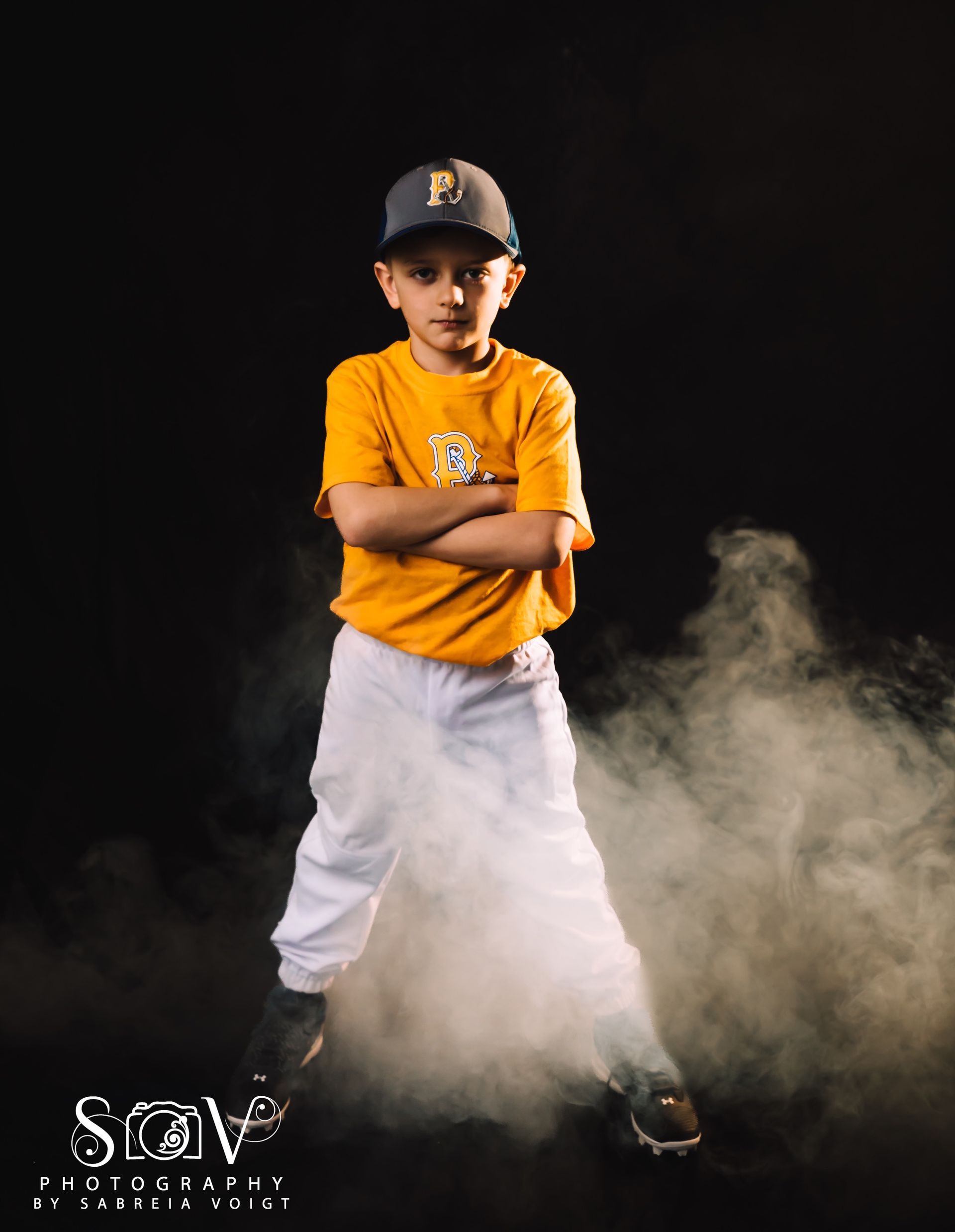 Boy in baseball uniform with arms crossed, standing in smoke against black background.