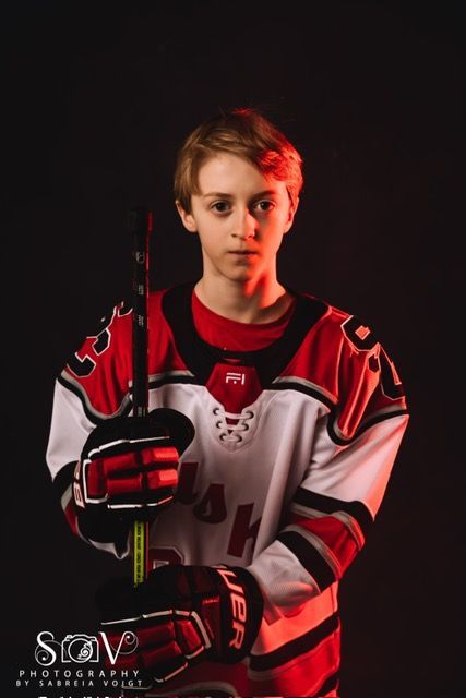 Young hockey player in a red and white jersey, holding a hockey stick. Dark background with red lighting.