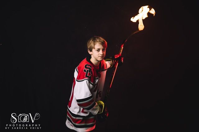 Boy in hockey jersey holds flaming hockey stick, dark background.