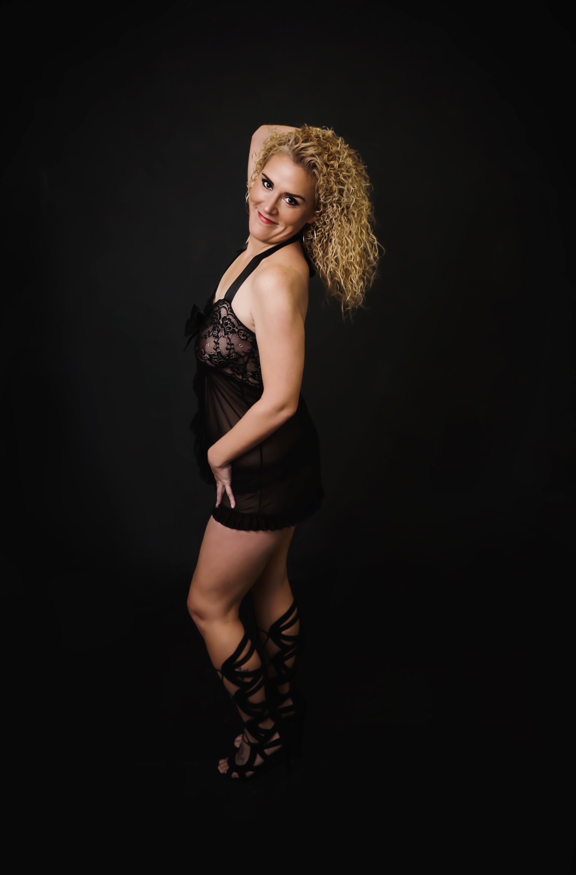 Woman in black lingerie and gladiator sandals, smiling, hand in hair. Dark background.