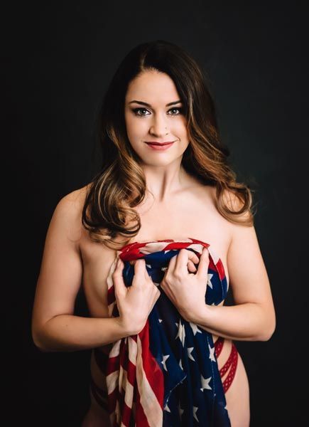 Woman, wrapped in American flag, smiling, dark background.