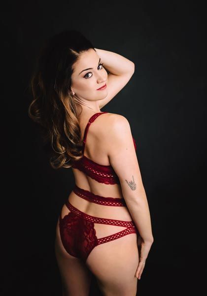 Woman in red lingerie, posing with hand on head, looking back, against a black backdrop.