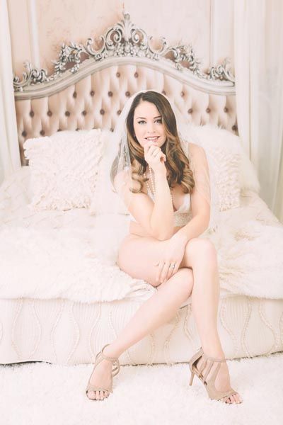 Woman in beige lingerie sits on bed, smiling, wearing heels.