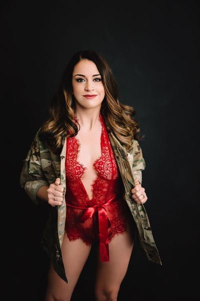 Woman in red lace bodysuit and camouflage jacket, smiling, posing in front of a black backdrop.